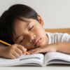 Close-up mata anak Asia yang mengantuk di meja belajar, dengan buku terbuka dan tangan memegang pensel.