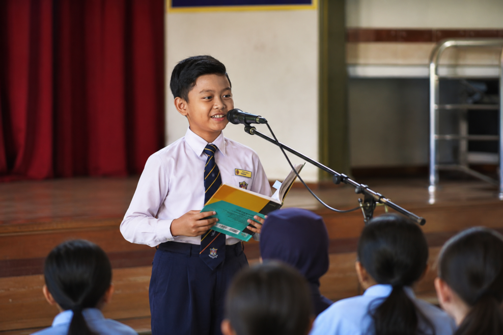 Kanak-kanak membaca dengan yakin di hadapan rakan sekelas dalam suasana cerah
