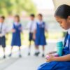 Anak Asia berseragam sekolah duduk sendirian di bangku semasa waktu rehat sambil memerhati rakan lain bermain dari jauh.