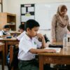 Murid Asia berseragam sekolah kelihatan murung apabila melihat kawannya dipuji, cikgu dan rakan lain di latar belakang.