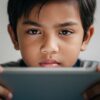 Close-up wajah anak Asia dengan mata letih dan fokus tegang sambil memegang telefon/tablet, suasana rumah tenang.
