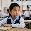 Close-up wajah murid Asia berseragam sekolah yang kelihatan malu, berhati-hati dan memandang ke arah rakan di luar bingkai.