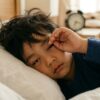 Close-up wajah anak Asia yang berat mata dan masih mengantuk pada waktu pagi, dengan jam/alarm kabur di latar belakang.