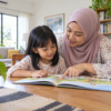 Ibu membimbing anak membaca buku asas dengan sabar.